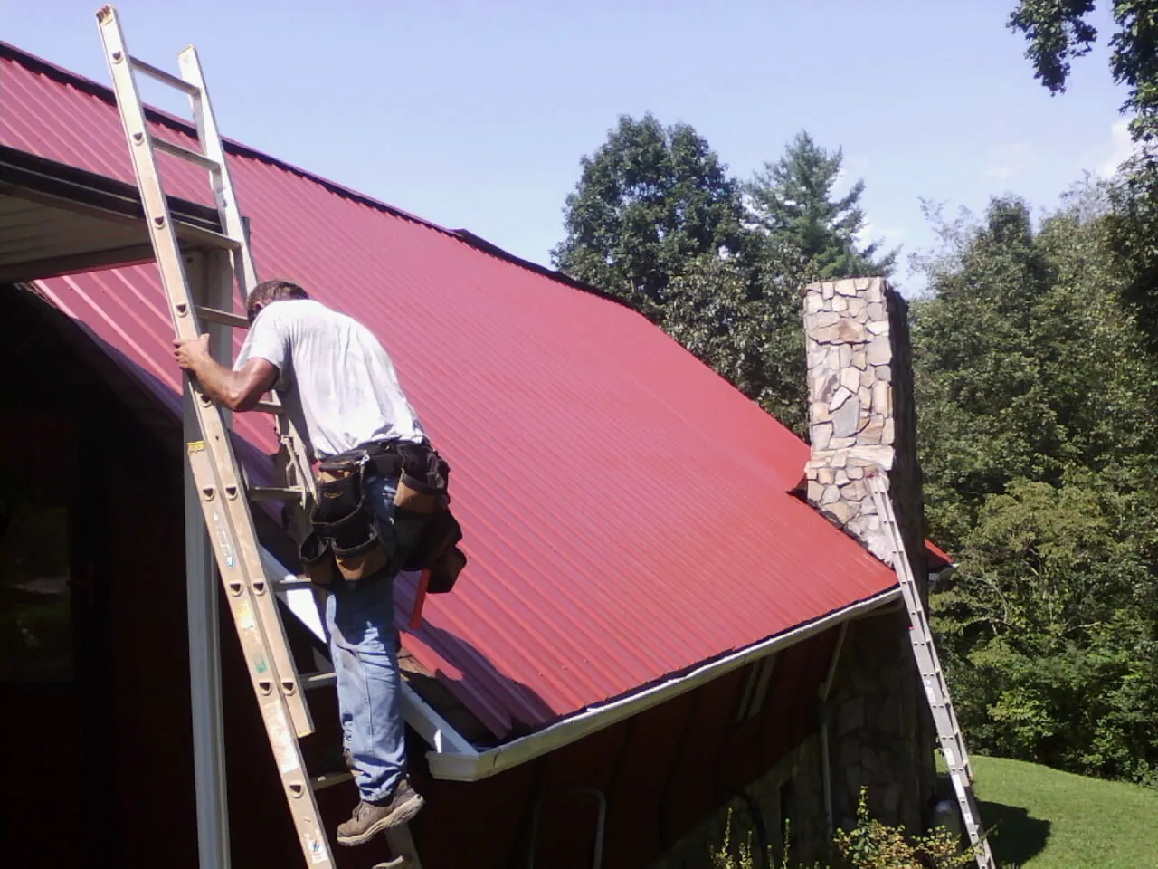Roofer on a ladder beside a red metal roof and stone chimney in Swedesboro